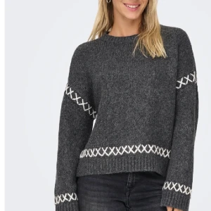 Mörkgrå stickad tröja med vita detaljer - Snygg mörkgrå stickad tröja med vita korsade detaljer runt ärmar, midja och handleder. Tröjan har en loose passform och långa ärmar, perfekt för att hålla sig varm och se chill ut. Ribbad kant vid hals, ärmslut och nederkant.