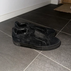 Axel Arigato Svarta slip-on sneakers i mocka  - Säljer ett par helsvarta slip-on sneakers i mocka med platt sula och rund tå. Skorna har en stilren och minimalistisk design utan snörning, vilket gör dem enkla att ta på och av. Perfekta för dig som gillar en clean look.