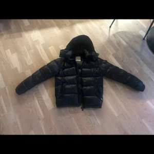 Svart Moncler dunjacka med huva - Svart dunjacka från Moncler med huva och klassisk logga på ärmen. Jackan har quiltad design, två dragkedjeförsedda fickor och en hög krage. Materialet är glansigt och fyllt med certifierat dun enligt DIST. Perfekt för kalla dagar och har en riktigt snygg streetstil.