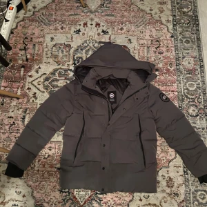 Mörkgrå Canada Goose Jacka - Säljer en mörkgrå dunjacka från Canada Goose med huva och snyggt broderat logomärke på ärmen.  Jag säljer den för att den inte passar längre och ej är min stil 