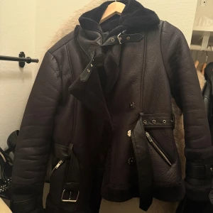 Svart pälsjacka från River Island - Säljer en svart pälsjacka från River Island med snygga detaljer som bälte, dragkedjor och spännen. Jackan har en fluffig krage och insida, samt flera coola metalldetaljer. Perfekt för dig som vill ha en varm och stilren look i vinter.