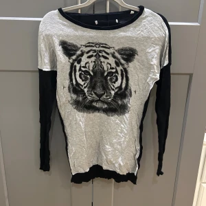 Svart och grå långärmad tröja med tiger - Cool långärmad tröja med svart och grå färgblock och ett stort tigertryck på framsidan. Tröjan har rund hals och är tillverkad i ett mjukt material som känns skönt mot huden. Perfekt för dig som gillar djurmotiv och vill sticka ut lite extra.