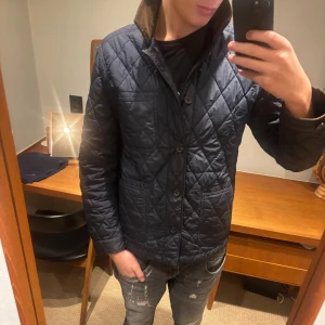 Polo Ralph Lauren jacka - Quiltad mörkblå jacka från Polo Ralph Lauren med bruna manchesterdetaljer på kragen. Jackan har knappar framtill, tre fickor och ett Polo-emblem på ärmen. Perfekt för höst och vinter, snygg och klassisk stil med loose passform.