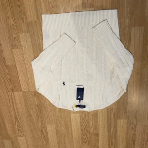 Vit kabelstickad tröja från Polo Ralph Lauren - Kabelstickad vit tröja från Polo Ralph Lauren med klassisk rund hals och broderad marinblå logga på bröstet. Tröjan har långa ärmar och breda ribbade muddar vid ärmslut och nederkant. Perfekt för en clean och stilren look.