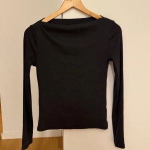 Mörkgrå långärmad topp - Säljer en långärmad topp med ribbad struktur och bred, rak halsringning. Modellen är slim och sitter snyggt mot kroppen. Perfekt att styla med jeans eller kjol för en clean look. Materialet känns mjukt och stretchigt, troligen bomull eller syntet.