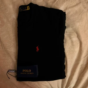 Svart t-shirt från Polo Ralph Lauren - Svart t-shirt från Polo Ralph Lauren med klassisk röd broderad logga på bröstet. Modellen är kortärmad och har en rund hals. Materialet är mjuk bomull som känns skönt mot huden. Perfekt för dig som gillar stilrena och tidlösa plagg med en exklusiv touch, Helt ny använd 1-2 gånger 