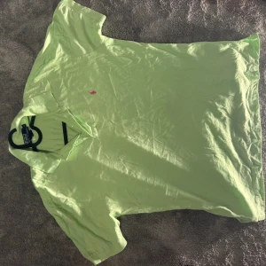 Ljusgrön Polo t-shirt från Ralph Lauren - Fräsch ljusgrön t-shirt från Polo Ralph Lauren i storlek XL. Klassisk pikékrage och korta ärmar, med den ikoniska rosa broderade loggan på bröstet. Tillverkad i mjuk bomull som är skön mot huden. Perfekt för dig som gillar stilren och enkel design.