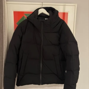 Uniqlo jacka - Säljer en svart pufferjacka med stor huva och dragkedja framtill. Jackan har en clean och stilren look med quiltad design och är perfekt för kalla dagar. Passformen är normal och jackan är långärmad. Enkelt att matcha med olika outfits.