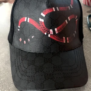 Svart Gucci keps med röd orm - Snygg svart keps från Gucci med diskret GG-mönster och en fet röd orm på framsidan. Kepsen har mesh på baksidan och tydliga Gucci-loggor på insidan. Materialet är polyester och syntet, perfekt för dig som vill sticka ut.