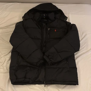 Svart pufferjacka från Polo Ralph Lauren - Svart pufferjacka från Polo Ralph Lauren med huva och röd broderad logga på bröstet. Jackan har flera dragkedjefickor, elastiska muddar och en stilren, oversized look. Perfekt för kalla dagar med sin tjocka vaddering och klassiska design.