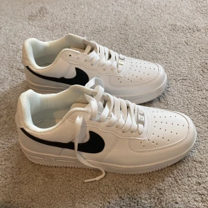 Nike Air Force 1 vit/svart sneakers - Fräscha Nike Air Force 1 sneakers i klassisk vit färg med svart swoosh. Lågt skaft, perforerad tå och platt sula för skön komfort. Skorna har snörning och är tillverkade i skinn med syntetdetaljer. Perfekt för dig som gillar stilrena och tidlösa sneakers.