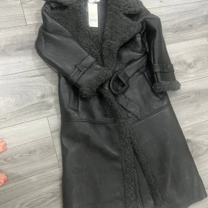 Adoore - Manchester coat - Säljer en svart lång teddyrock från Adoore med bred krage och bälte i midjan. Jackan har snygga detaljer i teddy på krage och ärmslut, samt ett coolt skinnliknande yttertyg. Perfekt för dig som vill ha en statementjacka med både stil och värme.