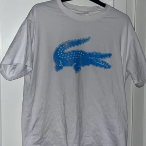 Vit Lacoste t-shirt - Vit t-shirt från Lacoste i bomull med stor blå krokodil-logga på bröstet. Loggan är lite sliten, därav skick 6/10. Storlek M, passar M