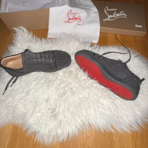 Christian Louboutin grå sneakers med nitar - Säljer ett par gråa sneakers från Christian Louboutin med ikoniska röda sulor och coola svarta nitar på tårna. Skorna är i mocka med snörning och har en rund tå. Perfekt för dig som vill sticka ut med lyxig streetstyle.