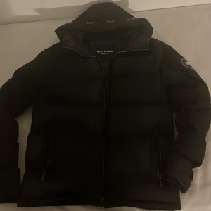 Svart pufferjacka Supply & Demand S - Svart pufferjacka från Supply & Demand med huva och snyggt emblem på ärmen. Jackan har dragkedja framtill och två fickor med dragkedja. Den är vadderad och har en modern, clean look som passar perfekt när det är kallt ute.