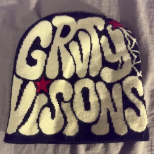 Svart mössa med tryck från Groovy Visions - Cool svart mössa med vit text 'Groovy Visions' i retrostil och röda stjärnor på framsidan. Baksidan har ett vitt spindelnätsmönster. Perfekt för dig som gillar unika detaljer och streetwear-vibe. Materialet är stickat och mjukt.