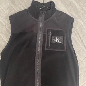 Svart fleeceväst från Calvin Klein - Svart fleeceväst från Calvin Klein Jeans med hög krage och hel dragkedja framtill. Västen har en praktisk bröstficka med dragkedja och CK-logga. Perfekt för lager-på-lager och chill stil.