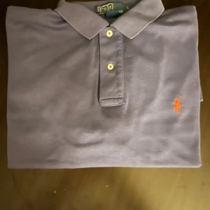 Lila pikétröja från Polo Ralph Lauren - Snygg lila pikétröja från Polo Ralph Lauren i 100% bomull. Klassisk krage, tre knappar framtill och orange broderad logga på bröstet. Perfekt för dig som gillar stilrena och ikoniska plagg med en twist av färg.