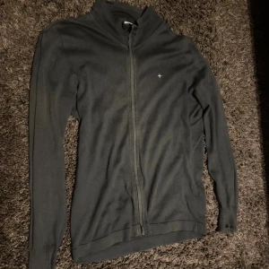 Svart kofta från Stone Island - Svart kofta från Stone Island med hel dragkedja framtill och diskret broderad logga på bröstet. Tröjan är tillverkad i mjuk bomull och har ribbade muddar vid ärmslut och nederkant. Perfekt för dig som gillar stilrena och klassiska plagg.