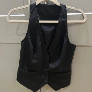 Svart kostym väst - Stilren svart väst från SHEIN i storlek 36. Klassisk modell med djup v-ringning, knappstängning framtill och två fickor. Västen har en rak passform och är tillverkad i ett slätt syntetmaterial som ger en clean look. Perfekt att styla över en t-shirt eller skjorta.