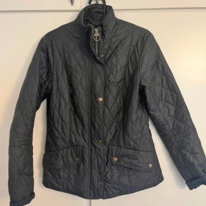 Svart quiltad Barbour jacka - Quiltad svart jacka från Barbour med ståkrage, guldfärgade knappar och dragkedja. Jackan har två fickor framtill med Barbour-logga och snyggt rutmönster över hela ytan. Perfekt för dig som gillar klassisk stil med modern touch.