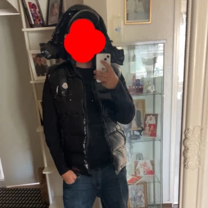 Moncler väst  - Snygg svart dunväst från Moncler med glansig finish och klassisk logga på bröstet. Västen har dragkedja framtill och en quiltad design som ger en cool och modern look. Perfekt att slänga över en hoodie eller t-shirt för extra stilpoäng. XL men passar även L