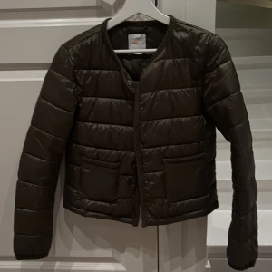 Mörk grön pufferjacka från Gina Tricot - Snygg mörk grön pufferjacka från Gina Tricot med rund hals och två framfickor med flätad kant. Jackan har tryckknappar framtill och är quiltad i horisontella sektioner. Perfekt för dig som gillar stilren och enkel design. Endast använd 2 ggr