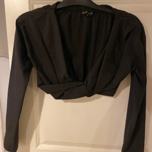 Svart croppad omlottopp från PrettyLittleThing - Säljer en svart croppad omlottopp från PrettyLittleThing. Toppen har lång ärm, djup v-ringning och elastisk rygg. Materialet är mjukt och lätt, perfekt för att styla med höga jeans eller kjol. Enkel och stilren design som ger en cool vibe.