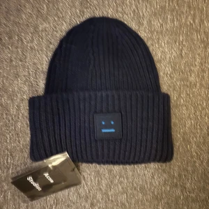 Mörkblå ribbstickad mössa Acne Studios - Snygg mörkblå ribbstickad mössa från Acne Studios med klassisk Face-logga framtill. Mössan är i mjuk ull och har bred uppvikt kant för extra stilpoäng. Perfekt accessoar för kalla dagar och en självklar favorit i garderoben.