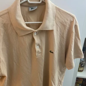 Beige pikétröja från Lacoste - Säljer en klassisk beige pikétröja från Lacoste med den ikoniska gröna krokodilloggan på bröstet. Tröjan har korta ärmar, krage och två knappar framtill. Perfekt för en clean och stilren look.