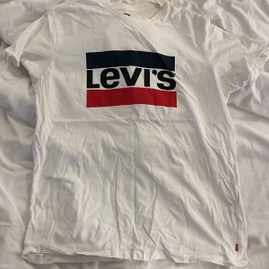 Vit Levi's t-shirt med logga, storlek L - Snygg vit t-shirt från Levi's i storlek L med klassisk stor logga framtill i svart, rött och mörkblått. T-shirten har rund hals och korta ärmar, tillverkad i mjuk bomull. Perfekt för dig som gillar streetwear och ikoniska märken.