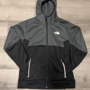 Svart och grå hoodie från The North Face - Säljer en snygg hoodie från The North Face i svart och mörkgrått. Tröjan har hel dragkedja, huva och två fickor med dragkedja. Perfekt för kyliga dagar och har en stilren logga på bröstet. Materialet är mjukt och känns sportigt.