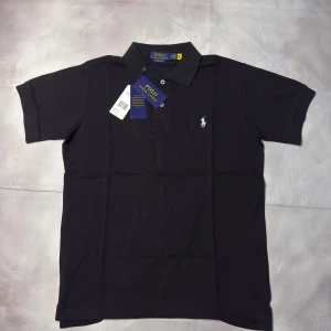 Svart pikétröja Polo Ralph Lauren - Svart pikétröja från Polo Ralph Lauren i bomull med klassisk krage och två knappar framtill. Slim fit-modell med korta ärmar och den ikoniska broderade loggan på bröstet. Perfekt för dig som gillar stilrena och tidlösa plagg.