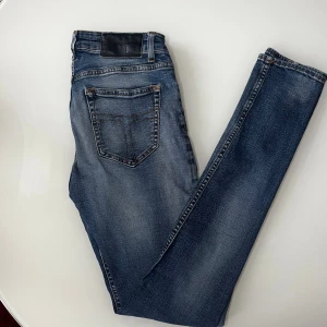 Tiger Of Sweden Jeans - Snygga blå jeans med klassisk femficksdesign och kontrastsömmar ifrån Tiger Of Sweden. Jeansen har en svart läderpatch bak i midjan och är tillverkade i slitstarkt denimtyg. Passformen är rak och de har en tidlös look som funkar till det mesta.
