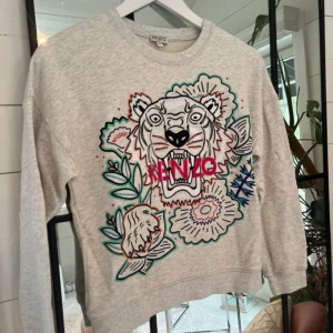 Grå sweatshirt med tiger från Kenzo - Säljer en grå sweatshirt från Kenzo med ett stort broderat tigerhuvud och färgglada blommor på framsidan. Tröjan har rund hals, lång ärm och normal passform. Kenzo-loggan är broderad i rosa framtill. Perfekt statement-plagg för dig som gillar coola prints.