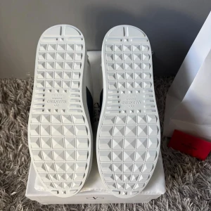 Valentino Garavani vita sneakers - Snygga vita sneakers från Valentino Garavani med marinblå detalj över snörningen. Skorna har rund tå, platt sula med geometriskt mönster och är tillverkade i skinn. Klassisk och stilren design med logga på plösen och innersulan. Perfekta för dig som gillar exklusiva märken.