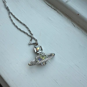 Vivienne Westwood halsband i silver - Unikt halsband från Vivienne Westwood i silverfärgad metall med ikonisk orb-berlock. Framsidan har glittriga stenar och en stjärna i mitten med regnbågsskimrande effekt. Kedjan är tunn och har ett coolt lås med logga. Perfekt statement piece för dig som gillar edgy stil. Vet inte om den är äkta köpt på vinted men hittar inga supés bara folk som säljer på eBay för 1000kr+