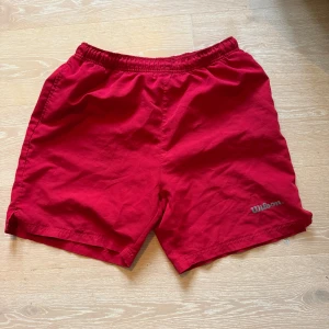Röda Wilson shorts med resårmidja - Snygga röda shorts från Wilson med elastisk midja och meshfoder. Shortsen har sidfickor och en bakficka med dragkedja, perfekta för sport eller chill. Klassisk Wilson-logga framtill. Lätta och bekväma att bära.