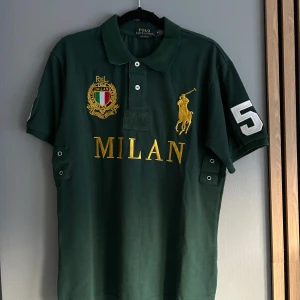 Grön pikétröja Polo Ralph Lauren Milan - Snygg grön pikétröja från Polo Ralph Lauren med stora broderade detaljer i guld och vitt, MILAN-text framtill och nummer 5 på ärm och rygg. Klassisk krage, korta ärmar och knappar vid halsen. Perfekt för dig som gillar sportig och stilren look.
