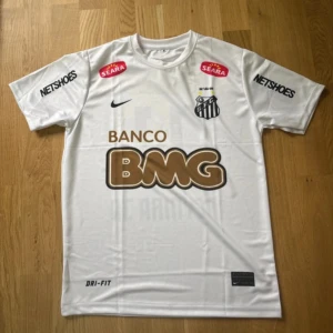 Vit Santos Neymar Jr fotbollströja - Säljer en vit Santos FC fotbollströja från Nike med Neymar Jr och nummer 11 på ryggen. Tröjan har korta ärmar, tryckta sponsorer och klubbmärke fram, samt BMG-logga i guld och svart. Materialet är lätt och andas, perfekt för match eller träning.