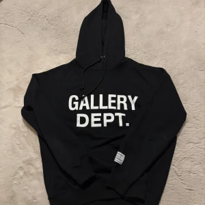Svart hoodie från Gallery Dept. - Svart hoodie från Gallery Dept. med stor vit logotyp på bröstet. Tröjan har huva med snörning och ribbade muddar vid ärmslut och nederkant. Materialet är mjuk bomull och passformen är normal. 