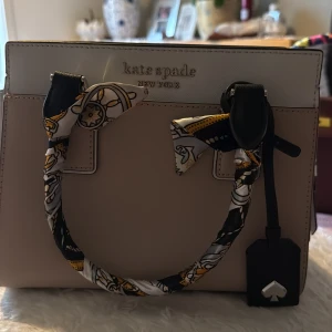 Beige handväska från Kate Spade - Snygg beige handväska från Kate Spade med stilren boxig form och dekorativa handtag insvepta i mönstrad scarf i svart, vitt och gult. Väskan har silverfärgade detaljer, innerfack med dragkedja och klassisk logga framtill. Perfekt för dig som gillar eleganta accessoarer.
