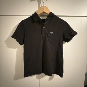 Svart pikétröja från Lacoste - Svart pikétröja från Lacoste med klassisk krage och korta ärmar. Tröjan har två knappar vid halsen och den ikoniska gröna krokodilloggan på bröstet. Tillverkad i mjuk bomull som är skön mot huden. Perfekt för dig som gillar stilren och sportig look.