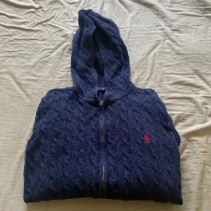 Marinblå stickad hoodie från Polo Ralph Lauren - Snygg marinblå stickad hoodie från Polo Ralph Lauren med dragkedja framtill och huva. Tröjan har kabelstickat mönster och en röd broderad logga på bröstet. Ribbstickade muddar vid ärmslut och nederkant. Perfekt för dig som gillar klassisk stil med en twist. Strl xl barn så passar s/m. Inga defekter. Använd gärna köp nu💕