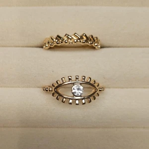 Guldiga ringar med ögonmotiv - Två snygga guldiga ringar, varav en har ett coolt ögonmotiv med en glittrande sten i mitten. Den andra ringen har en unik, taggig design som ger extra edge till din stil. Perfekta accessoarer för att sticka ut och addera lite bling till din look.
