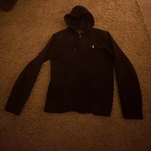 Mörkblå hoodie från Polo Ralph Lauren - Mörkblå hoodie från Polo Ralph Lauren med dragkedja och klassisk huva. Tröjan har lång ärm och den ikoniska lilla loggan broderad på bröstet. Perfekt för en chill och stilren look. Materialet är mjuk bomull som känns skönt mot huden. Pris kan diskuteras, nypris ca 2000