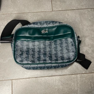 Mönstrad axelväska från Lacoste - Snygg axelväska från Lacoste i mörkgrönt och svart med geometriskt mönster i vitt. Väskan har justerbar svart axelrem, silverfärgad krokodillogo och dragkedja upptill. Perfekt storlek för att få med allt viktigt och stilren design.