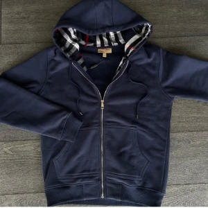 Marinblå Burberry hoodie med dragkedja - Säljer en marinblå hoodie från Burberry med klassiskt rutigt foder i huvan. Tröjan har dragkedja, snörning vid huvan och två fickor framtill. Tillverkad i mjuk bomull och har broderad Burberry-logga på bröstet. Perfekt för dig som gillar stilren och lyxig streetwear.