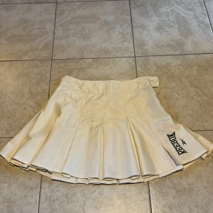 Vit plisserad kjol från Reebok XS - Superfräsch vit plisserad kjol från Reebok i sportig stil. Kjolen har bred linning, tydliga veck och svart Reebok-logga på ena sidan. Tillverkad i polyester, perfekt för tennis eller en aktiv look.