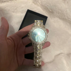 Glittrig guldig klocka med blå urtavla - Säljer en riktigt blingad klocka med guldig länk och massor av glittrande stenar. Urtavlan är ljusblå med glittriga siffror och visare i guld. Klockan har en rund form och ett lyxigt, iögonfallande utseende som verkligen sticker ut.